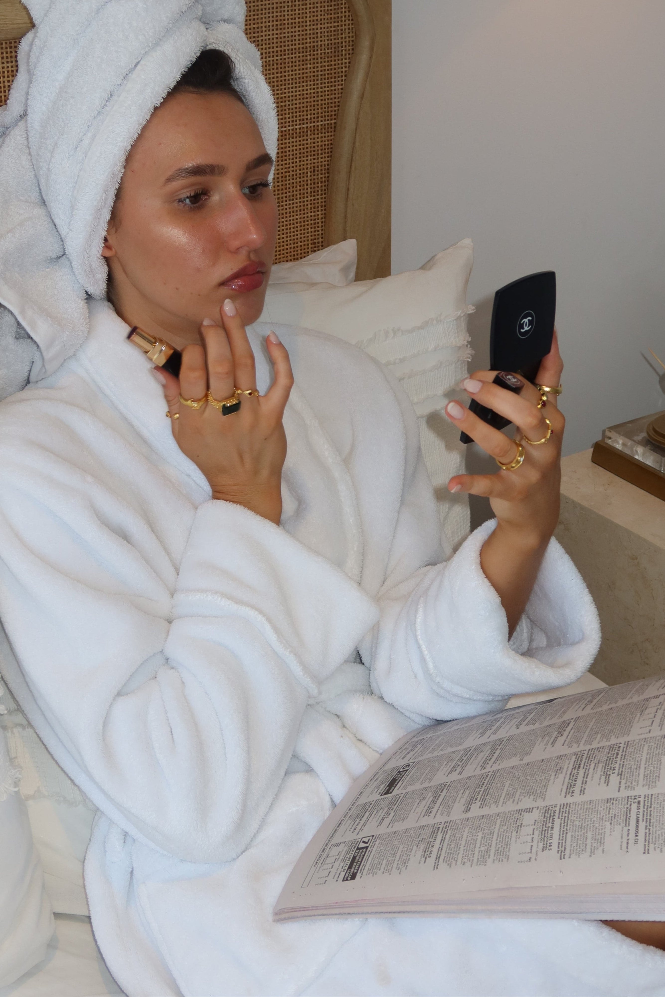 Person lying in bed with a towel on their head, holding a phone and reading a newspaper.